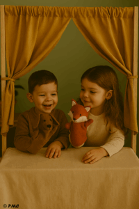 Deux enfants jouant avec des marionnettes chien et renard derrière un petit théâtre en tissu, dans une activité d’expression scénique.