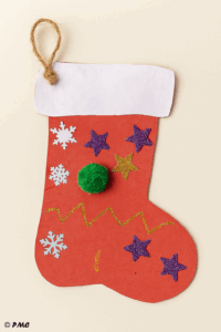 Guirlande de bottes de Noël décorées par des enfants, découpées dans du papier rouge et suspendues à une ficelle. Chaque botte est ornée de paillettes, pompons, gommettes et contient un petit souhait plié à l’intérieur.