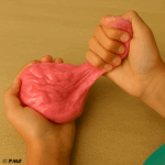 Préparation d’un slime rose nacré avec les ingrédients posés sur une table et la main d’un enfant versant du liquide