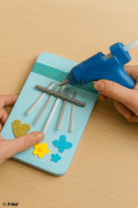 Mains d’un adulte fixant les tiges métalliques sur une kalimba artisanale avec un pistolet à colle.