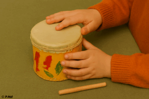 Mains d’un enfant jouant sur un tambour fait maison avec une membrane tendue sur une boîte décorée, accompagnée d’un petit bâton.