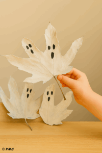 Feuilles peintes en fantômes suspendues à des branches pour un bricolage d’Halloween fait par un enfant