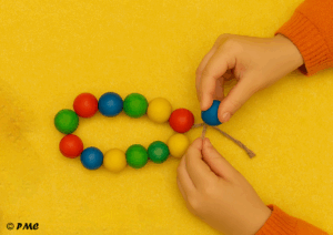 Mains d’un enfant en train d’enfiler des perles colorées sur une cordelette, dans le cadre d’une activité de motricité fine.