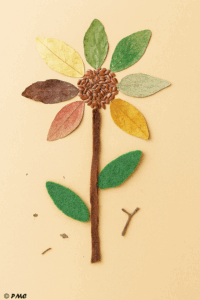 Fleur artisanale réalisée avec des feuilles séchées, graines de lin, tissu et feutrine, assemblée grossièrement par un enfant sur fond beige.