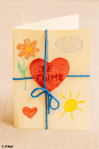 Carte pliée décorée par un enfant avec un cœur rouge, des dessins au crayon et un fil bleu noué