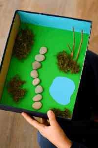 Diorama enfant avec chemin en cailloux, mousse pour les buissons et brindilles plantées comme arbres miniatures.