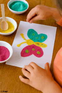 Main d’enfant saupoudrant du sable coloré sur les ailes d’un papillon encollé.