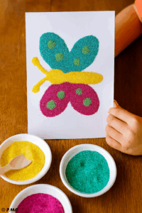 Tableau de sable coloré représentant un papillon réalisé par un enfant, avec du sable turquoise, magenta, jaune et vert.